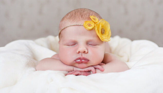 baby girl facing the camera laying on her tummy on a white blanket with yellow flower hairband