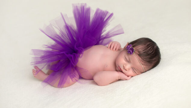 Sleeping, newborn baby girl wearing a purple, tulle tutu and flower headband.